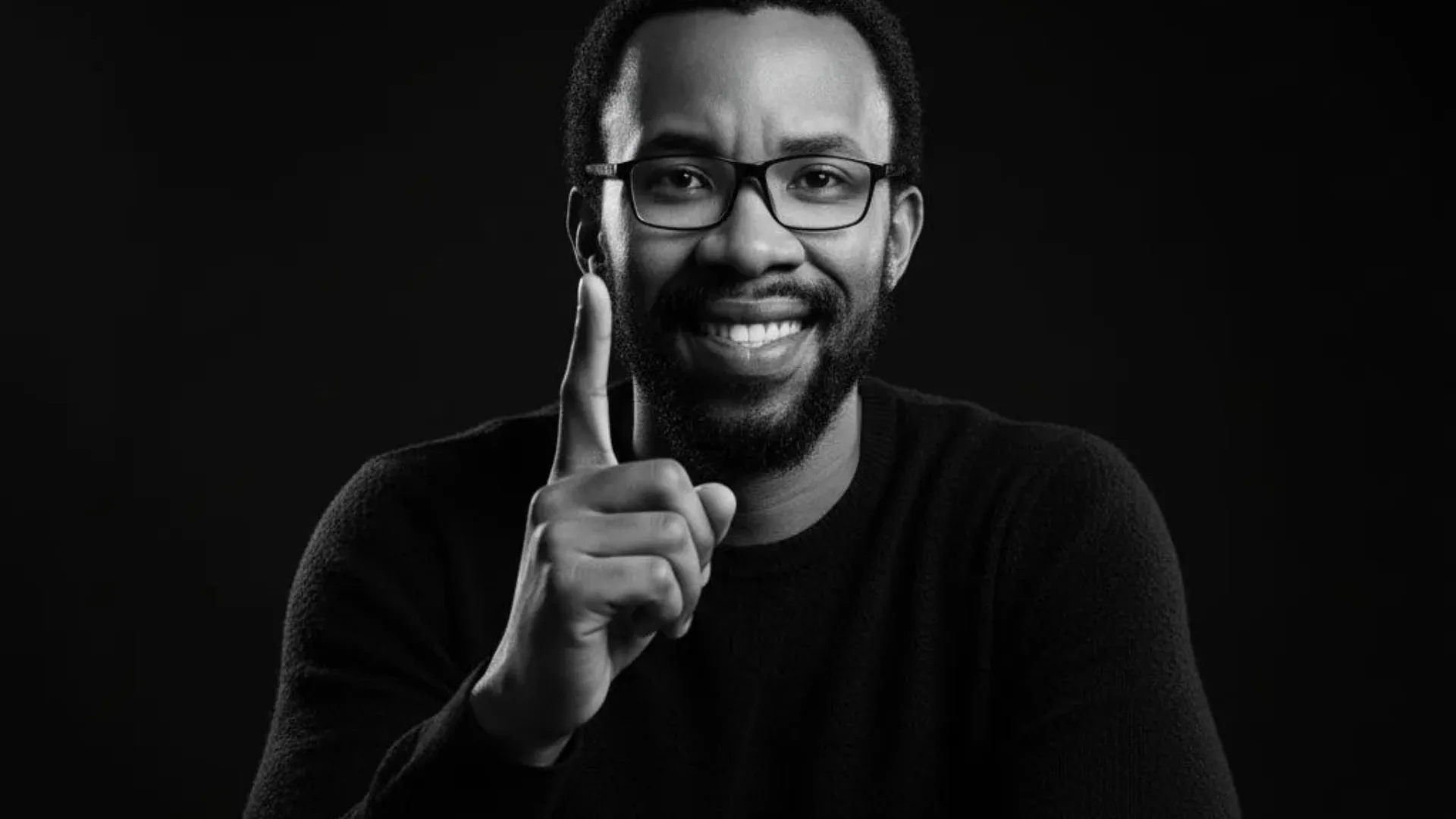 Black and white portrait of a smiling man with glasses and beard wearing a dark sweater against black background