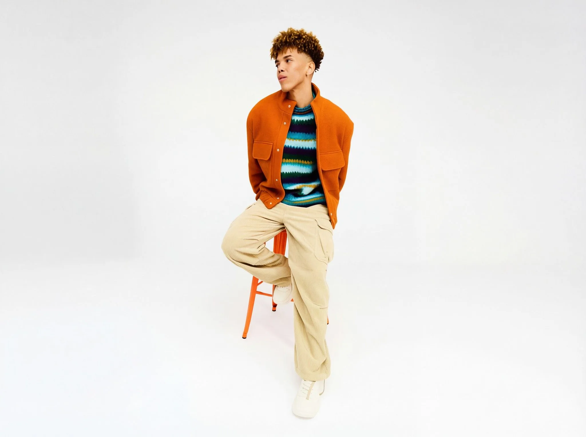 Young man with curly hair wearing bright orange jacket, blue striped shirt, and beige pants, sitting on orange chair against white background