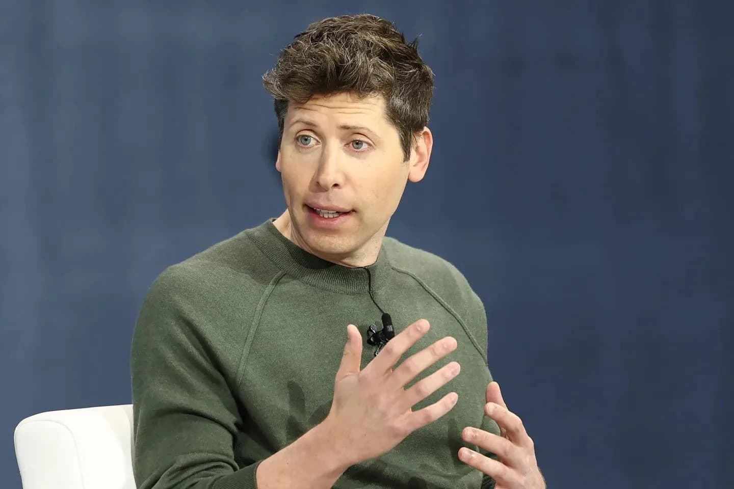 Man in olive green sweater speaking with hand gestures against blue background during interview or presentation