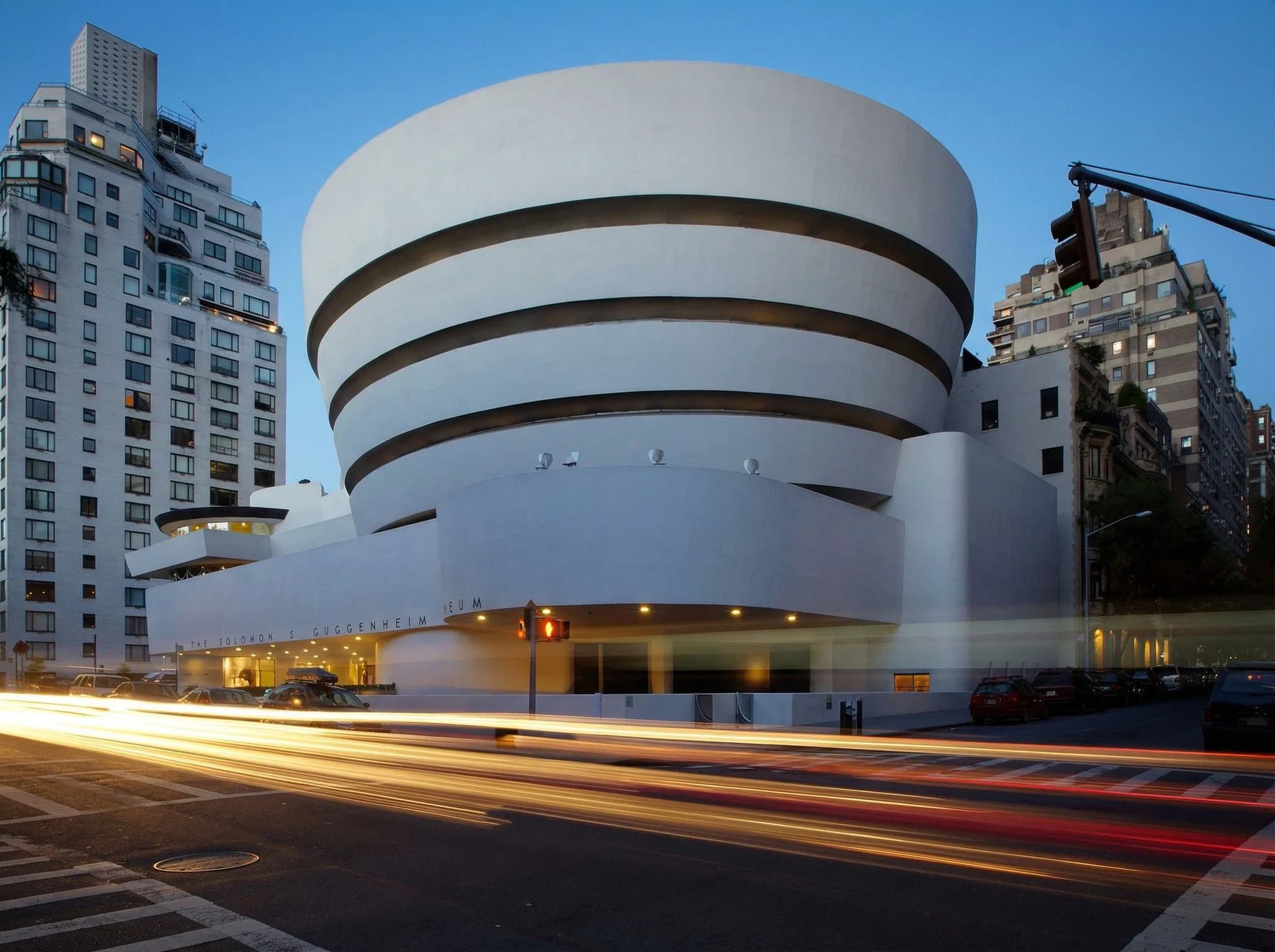 Guggenheim Museum at dusk with iconic spiral white exterior, Manhattan skyline, and light trails from traffic on 5th Avenue, NYC