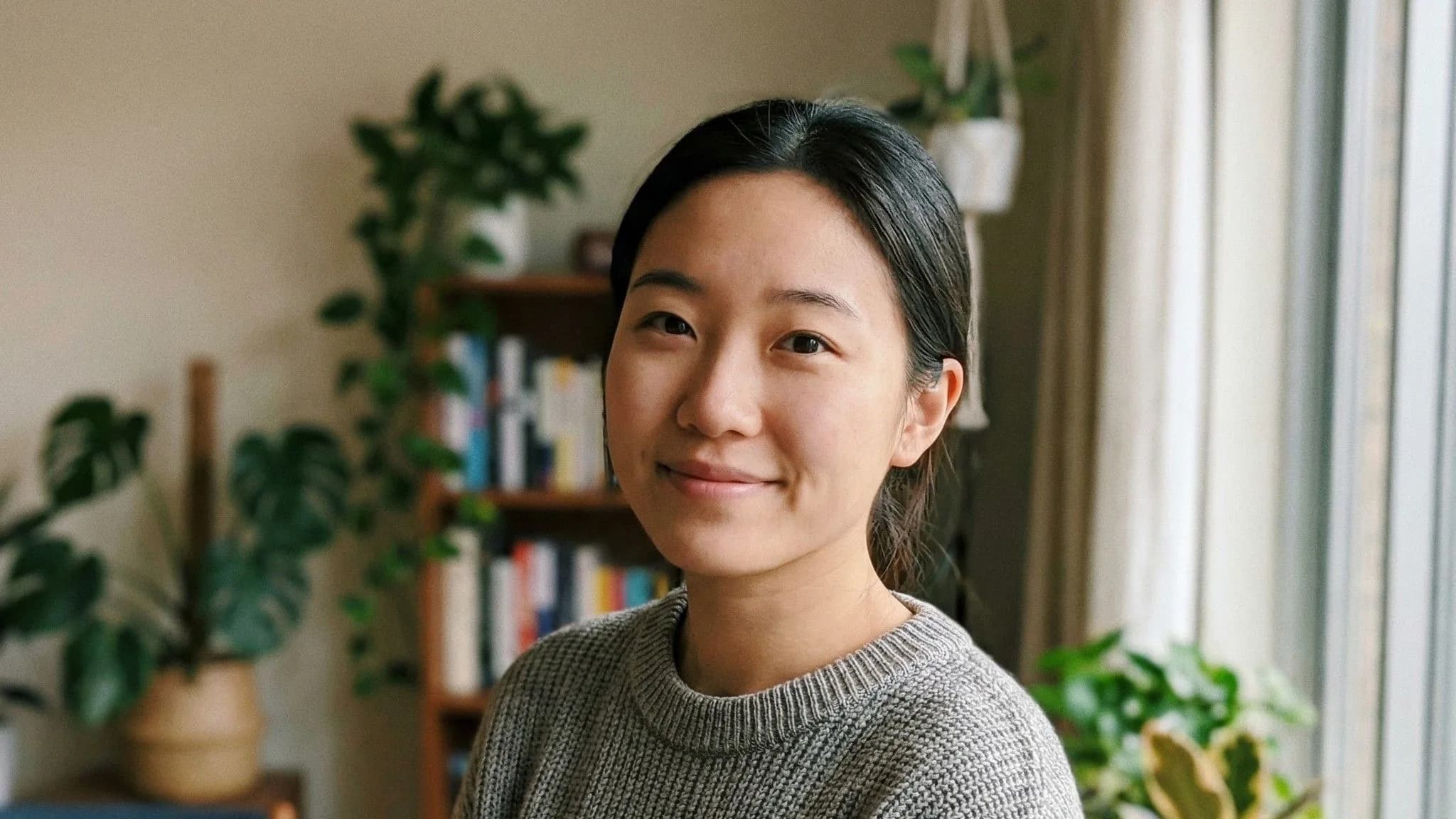 Portrait of smiling Asian woman in gray sweater indoors with bookshelves and potted plants