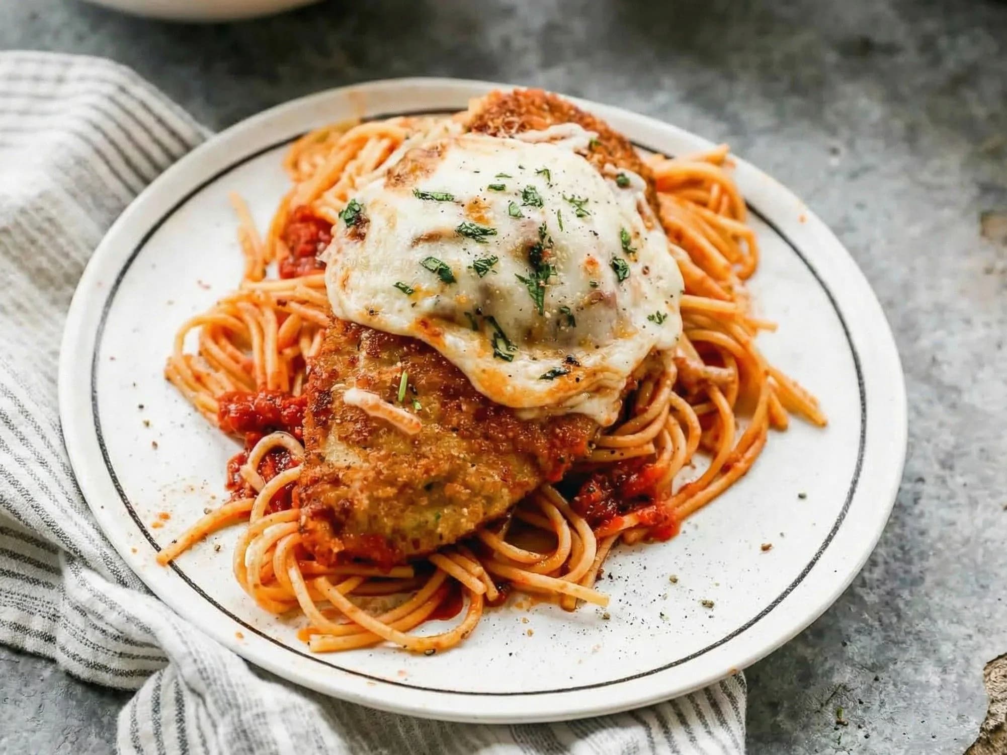 Chicken Parmesan with melted cheese on spaghetti in red tomato sauce, garnished with fresh parsley on a white plate