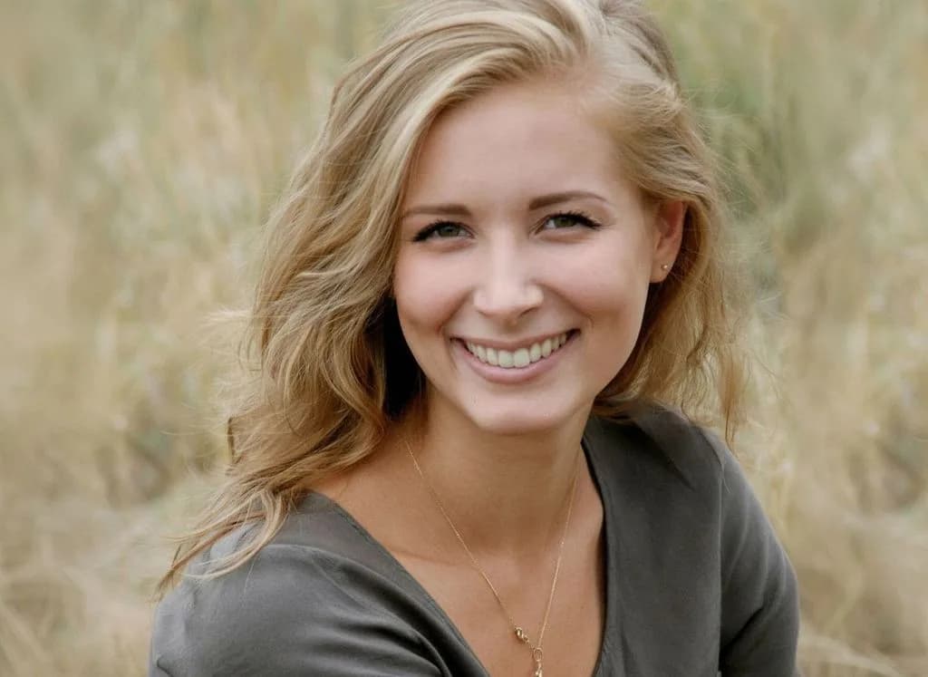 Smiling blonde woman wearing gray top with necklace in professional headshot portrait against neutral background