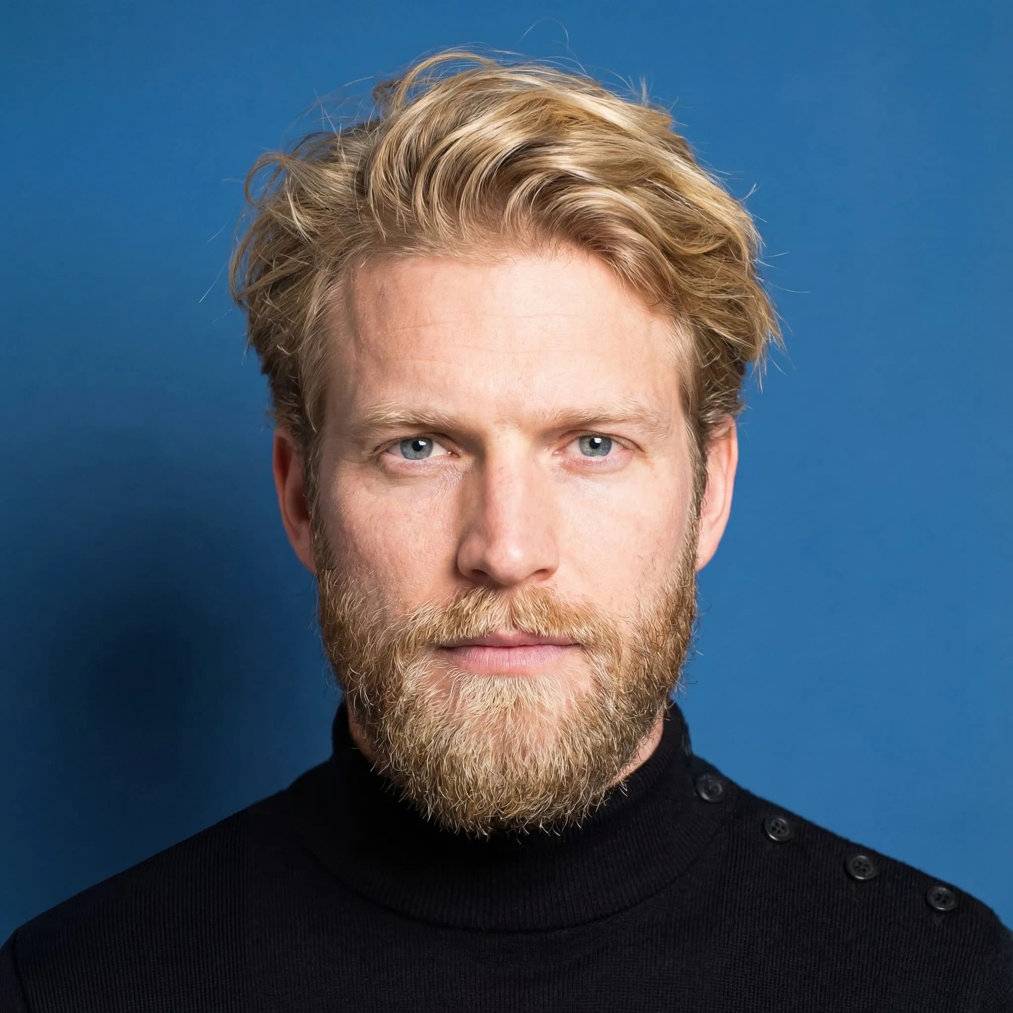 Portrait of man with blonde wavy hair and full beard wearing black turtleneck against blue background