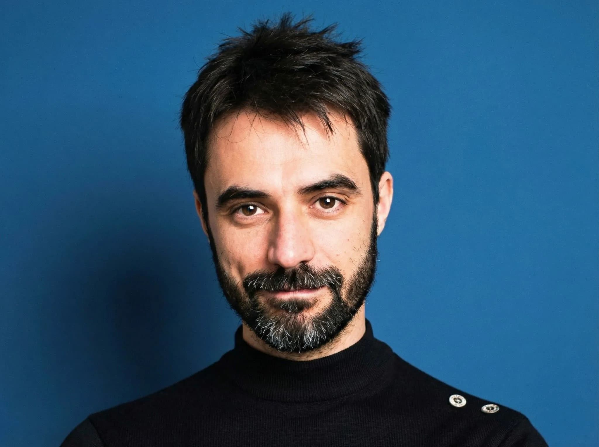 Bearded man with dark hair wearing black turtleneck against blue background professional headshot portrait