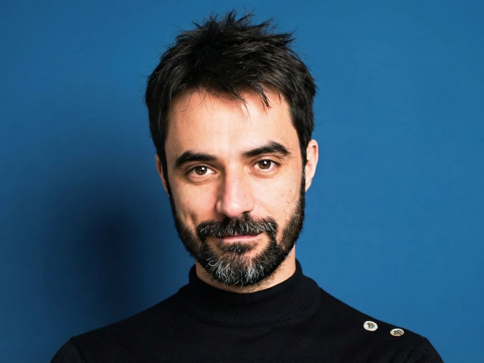 Portrait of bearded man with dark hair wearing black turtleneck sweater against solid blue background