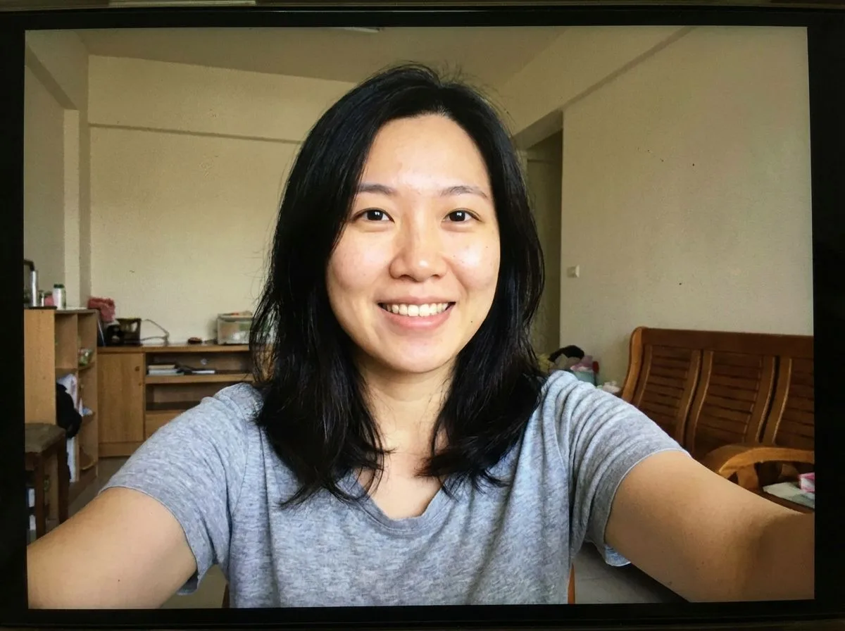 Smiling Asian woman with long black hair wearing gray t-shirt taking selfie in bedroom home interior