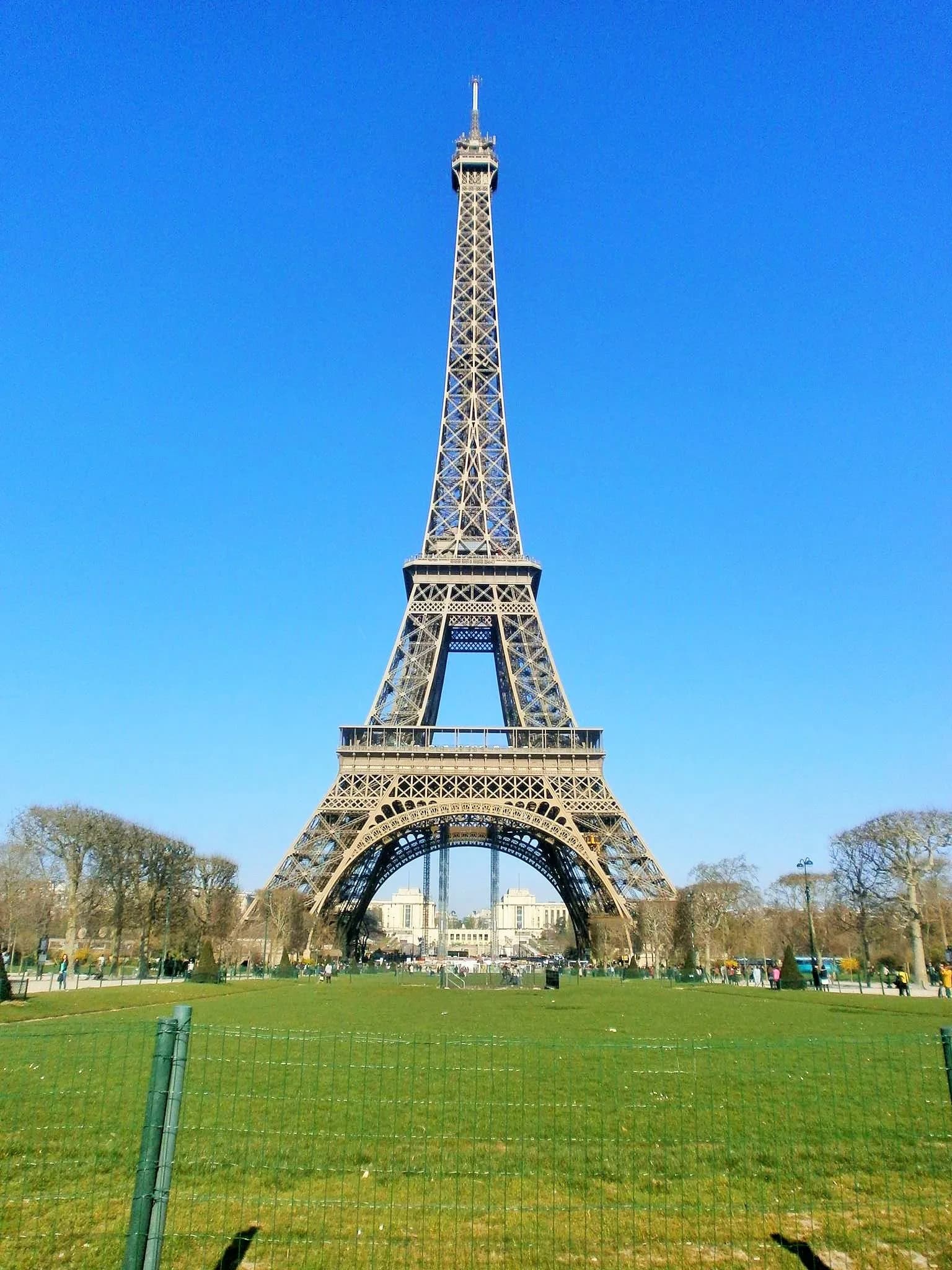 Eiffel Tower in Paris viewed from Champ de Mars lawn with clear blue sky and tourists, iconic French monument