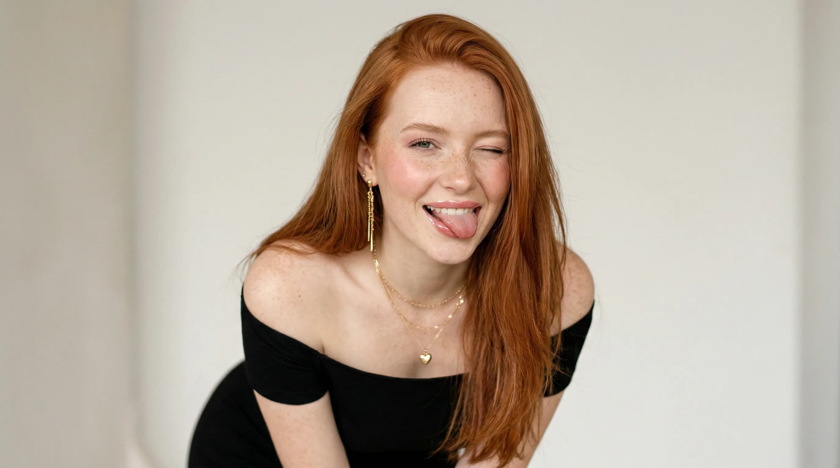 Playful young woman with long red hair wearing black off-shoulder top and gold jewelry, smiling with tongue out against white background