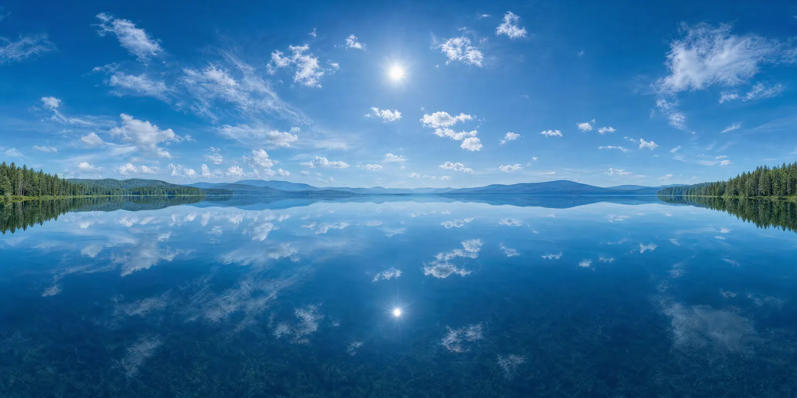 360 panorama of a calm lake at noon under a blue sky with white clouds and a forested shore