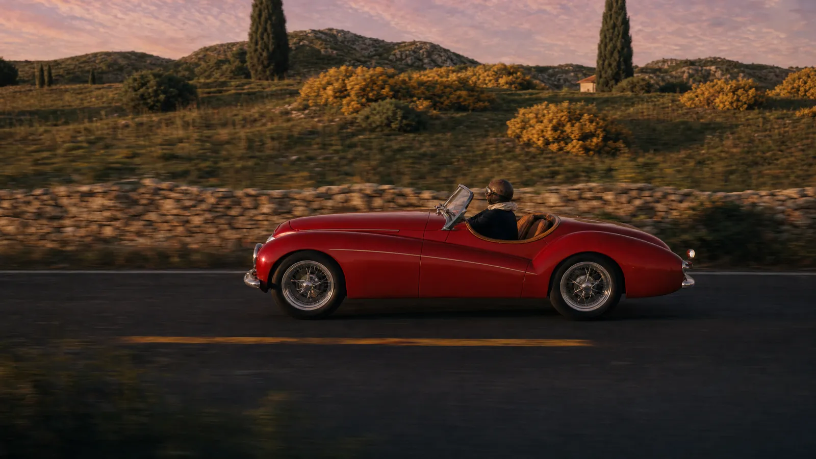 Style frame for the tracking: the roadster in profile on an asphalt road beside a dry-stone wall, gorse and cypresses in low golden light